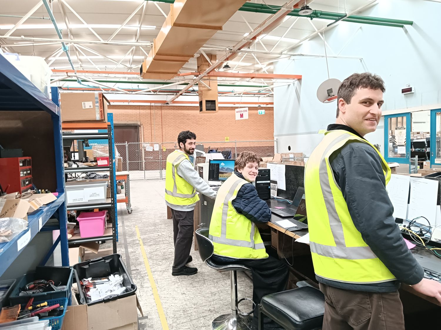Three volunteers in hi-vis vests smiling at the build room benches